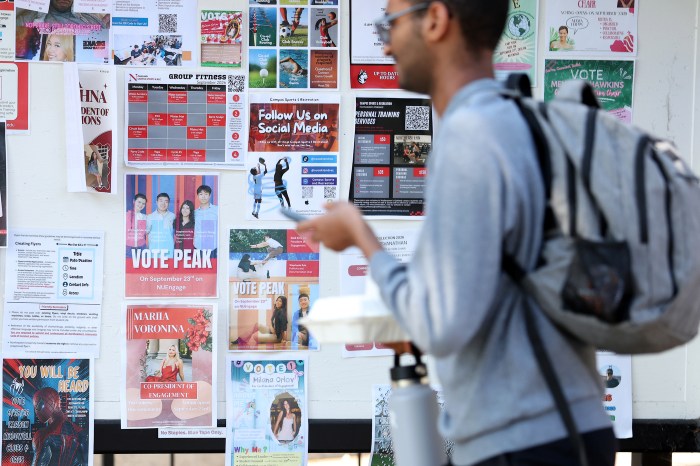 Student walks past a board covered in campaign flyers and posters on Northeastern's Oakland campus.