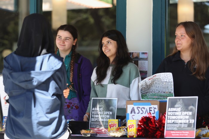Three students talk and smile together indoors, with campaign flyers visible on the windowsill behind them.