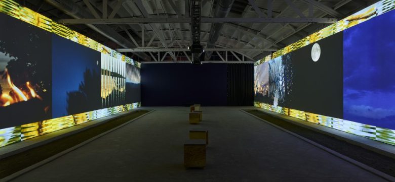 A dimly lit gallery features large digital screens on two walls, displaying images of fire, cityscape, and the moon, with wooden benches arranged in the center.