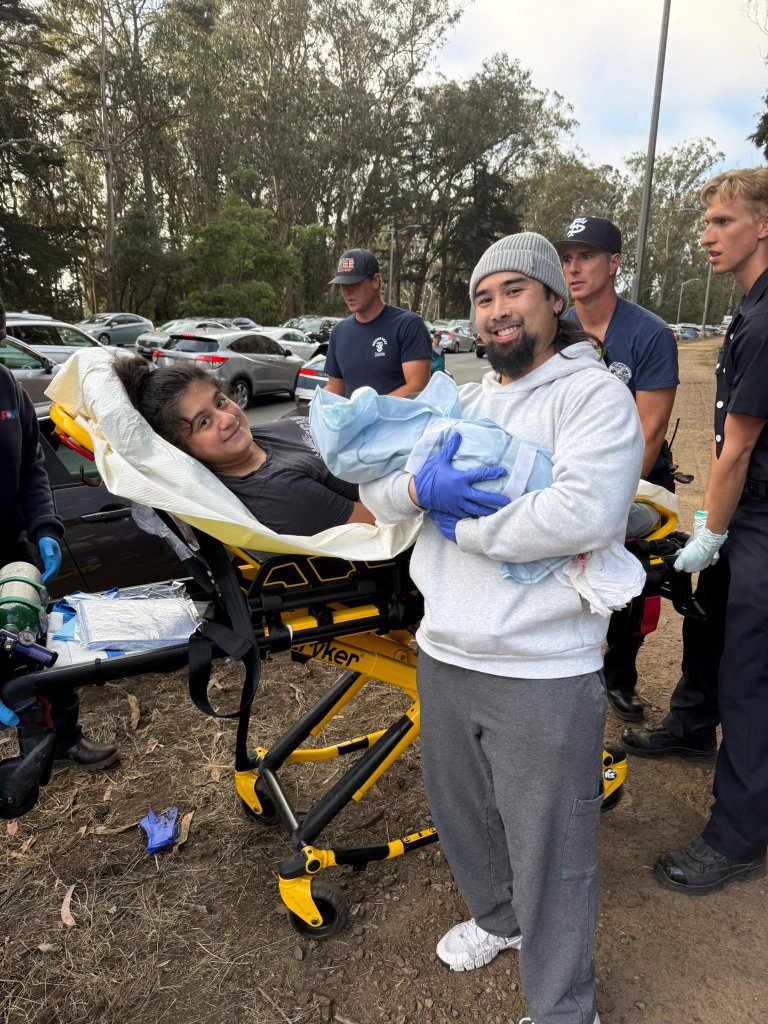 A man holds a newborn baby while standing next to a woman on a stretcher, with emergency responders in the background.