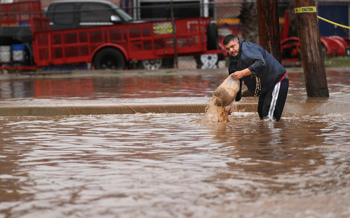 A man standing on a roadway in knee-deep muddy water tries to clear a drain.