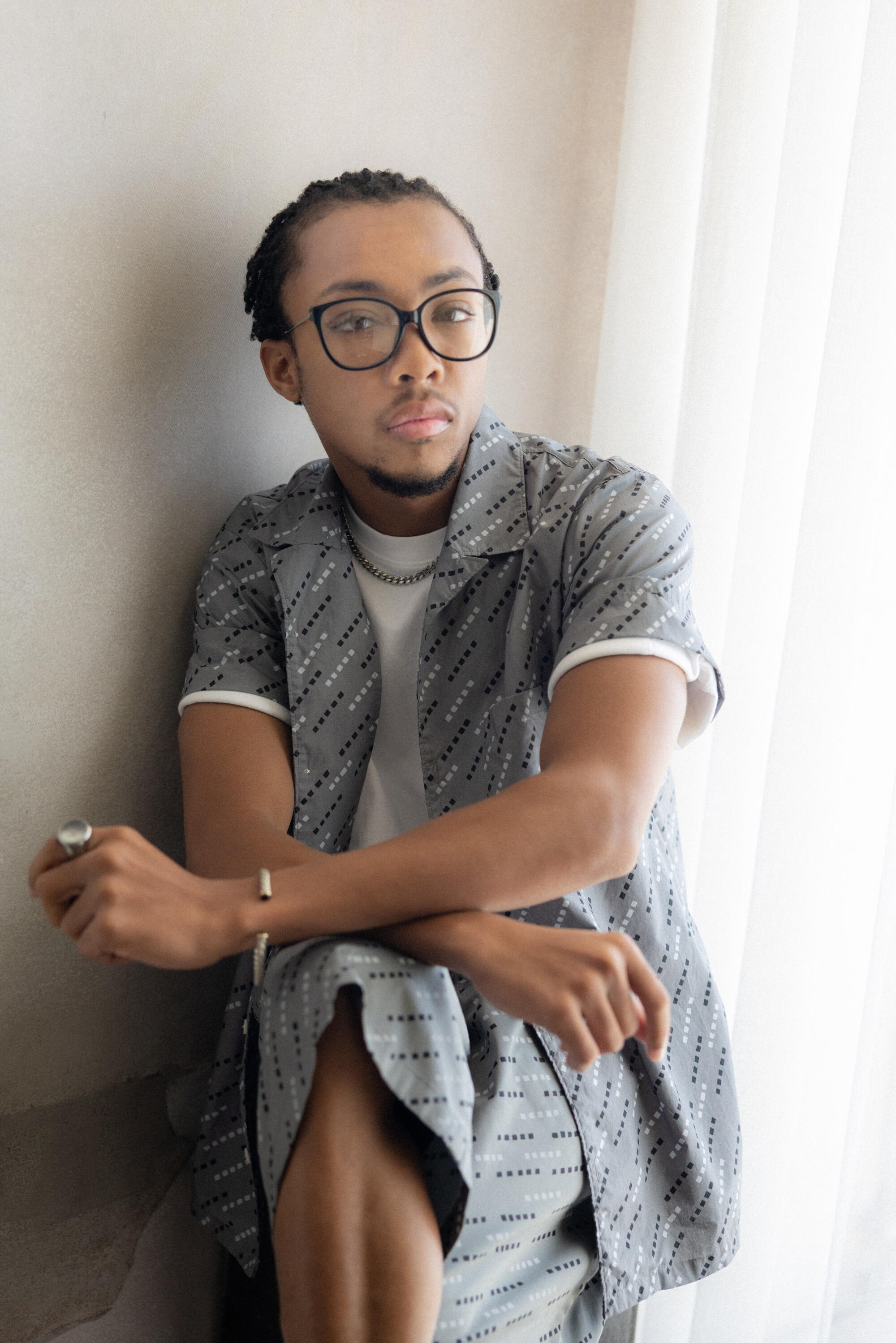 A man wearing glasses and blue top and shorts sits near a white wall.