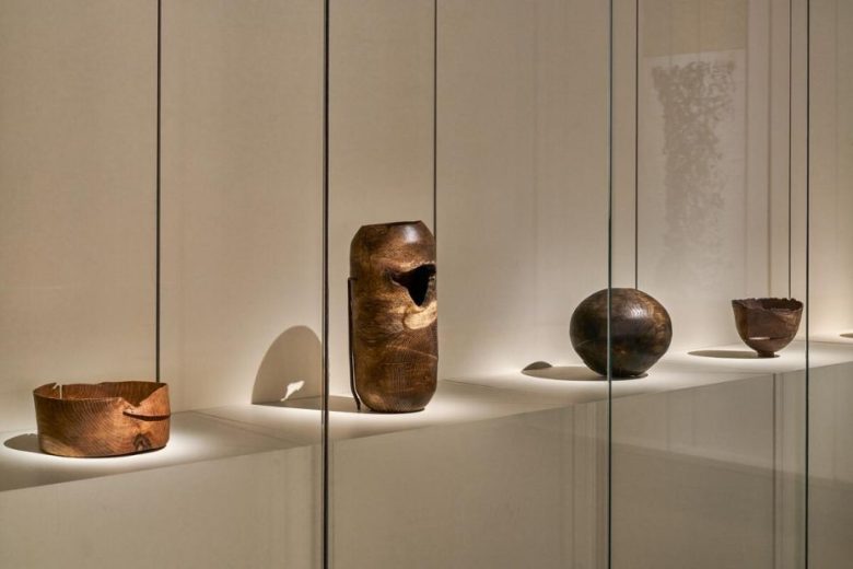 Five wooden vessels of varying shapes and sizes are displayed on a white shelf behind glass in a well-lit gallery setting.
