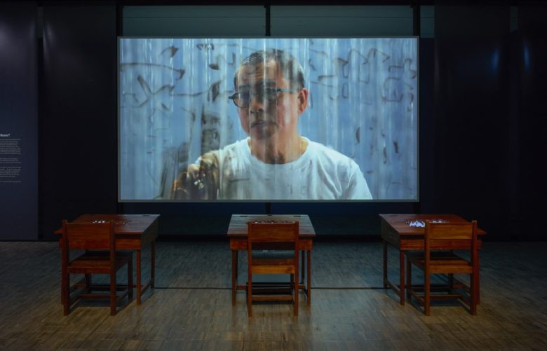 Large screen displays a man in glasses behind three wooden desks and chairs in a dimly lit room with dark walls and wooden flooring.