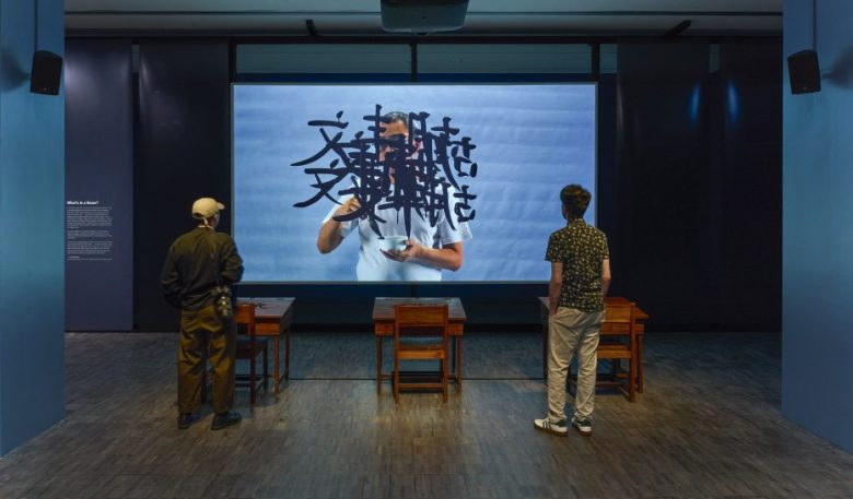 Two people stand facing a large screen displaying a person with black abstract symbols over their face, in a dimly lit room with two wooden desks.