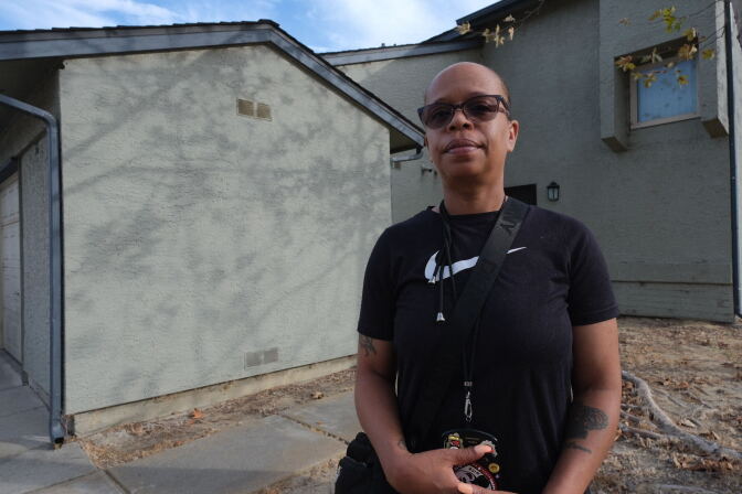 A woman with dark skin tone, wearing sunglasses and a black Nike t-shirt, stands outside the front door of townhome in San Pedro.