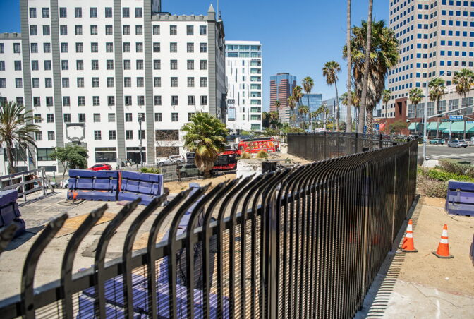 A black metal fence stretches down a street overlooking a cement area with blue barriers. Office buildings and palm trees are in the background.