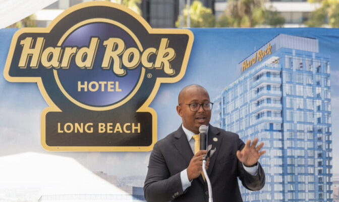 Mayor Rex Richardson, a man with dark skin tone, wearing a suit and tie, speaks into a handheld microphone and gestures with his hand as he stands in front of signage that has a rendition of a tall building and text that reads "Hard Rock Hotel. Long Beach."
