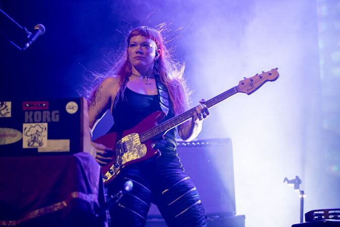 A musician plays an electric guitar on stage, hair blowing, surrounded by purple stage lighting and haze.