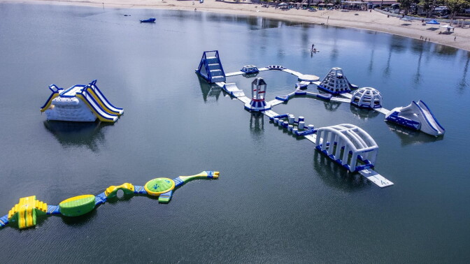 Inflatable water park with slides, climbing structures, and obstacle courses floats on a lake near a sandy beach.