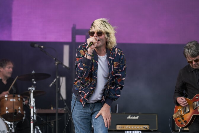 A singer with sunglasses and blond hair performs into a microphone on stage, with a drummer and guitarist nearby.