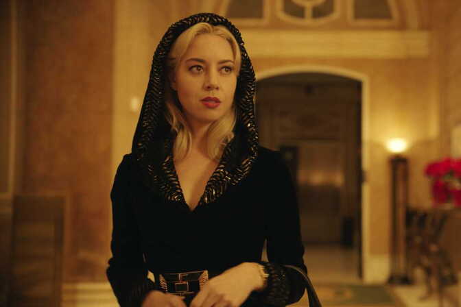 A blonde person in a black hooded coat with a jeweled belt stands in an ornate hallway.