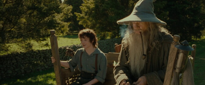 A person in a wide-brimmed hat smokes a long pipe beside a younger person on a wooden bench, trees and stone walls behind them.