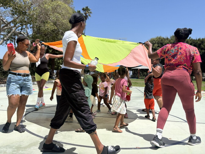 Black moms are walking in a circle holding a parachute while children are dancing underneath it.