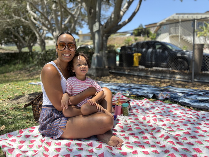 An adult holds a child on a watermelon-print picnic blanket under a tree, with cars and buildings in the background.