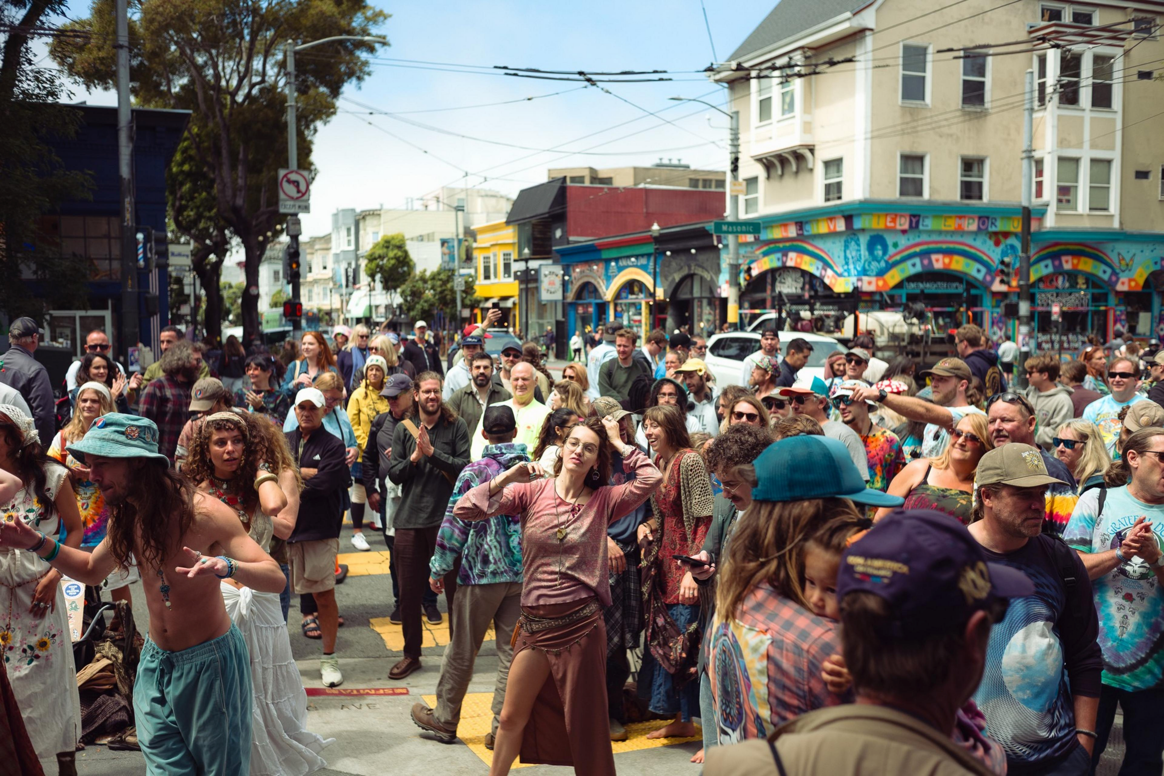 A diverse crowd dances and mingles outdoors on a sunny street near a colorful, mural-covered building. Many wear casual and tie-dye clothing.