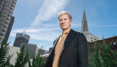A man in a black jacket stands outside, with skyscrapers and clear blue sky featuring wispy clouds in the background. Green foliage is in the foreground.