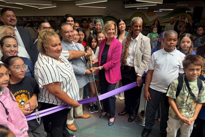 People hold giant scissors to cut a purple ribbon at a library ceremony, surrounded by children and adults.