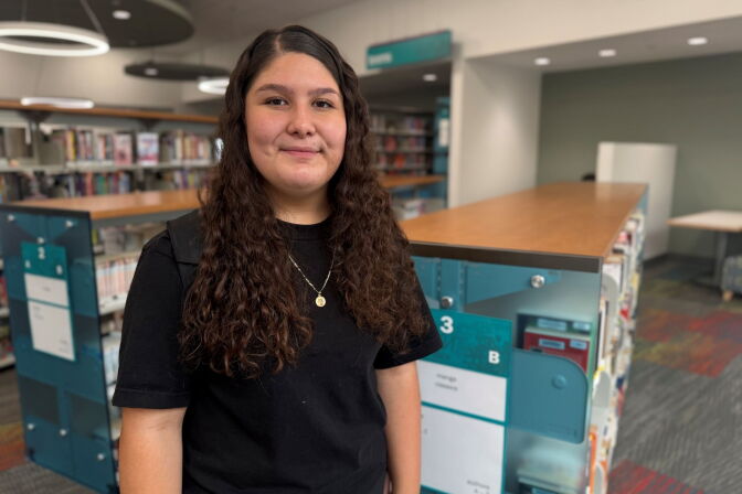 A girl with light skin tone and long curly hair and a black t-shirt stands smiling in a library near bookshelves.
