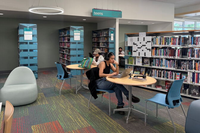 People work at tables in a library's teen section, surrounded by bookshelves.