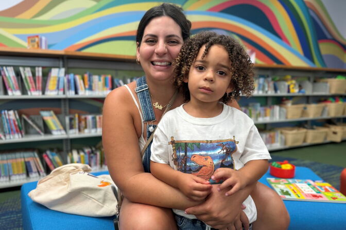 With library shelves in the background, a woman with medium-light skin tone holds a child with medium-skin tone and curly hair.