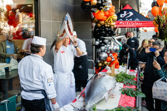 A man holds up a tuna head, as he goes through the process of cutting the fish, with an audience in attendance.