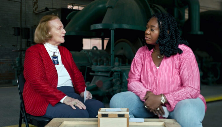 An older white woman at right, near 80 years old, speaks with a thoughtful expression while a younger Black woman, near 30, listens.