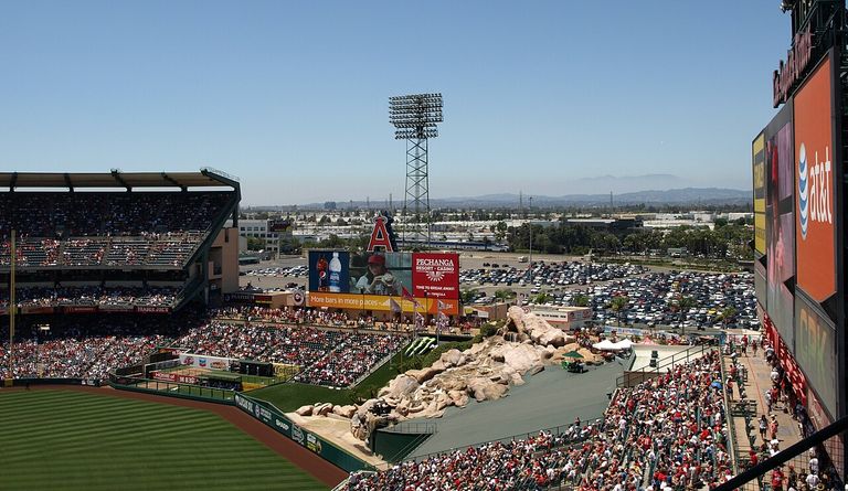 Angels baseball stadium with a packed crowd, large scoreboard, outfield rock feature, and distant view of mountains under a clear blue sky.