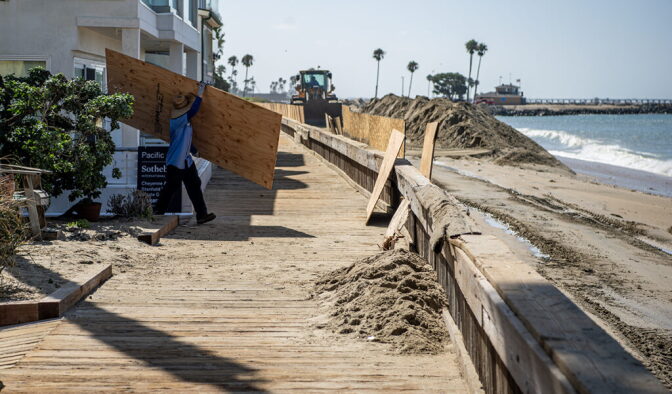 A person holds a plywood board over their shoulder walking towards a sea barrier on a boardwalk next to homes as a tractor on a beach pushes piles of sand.