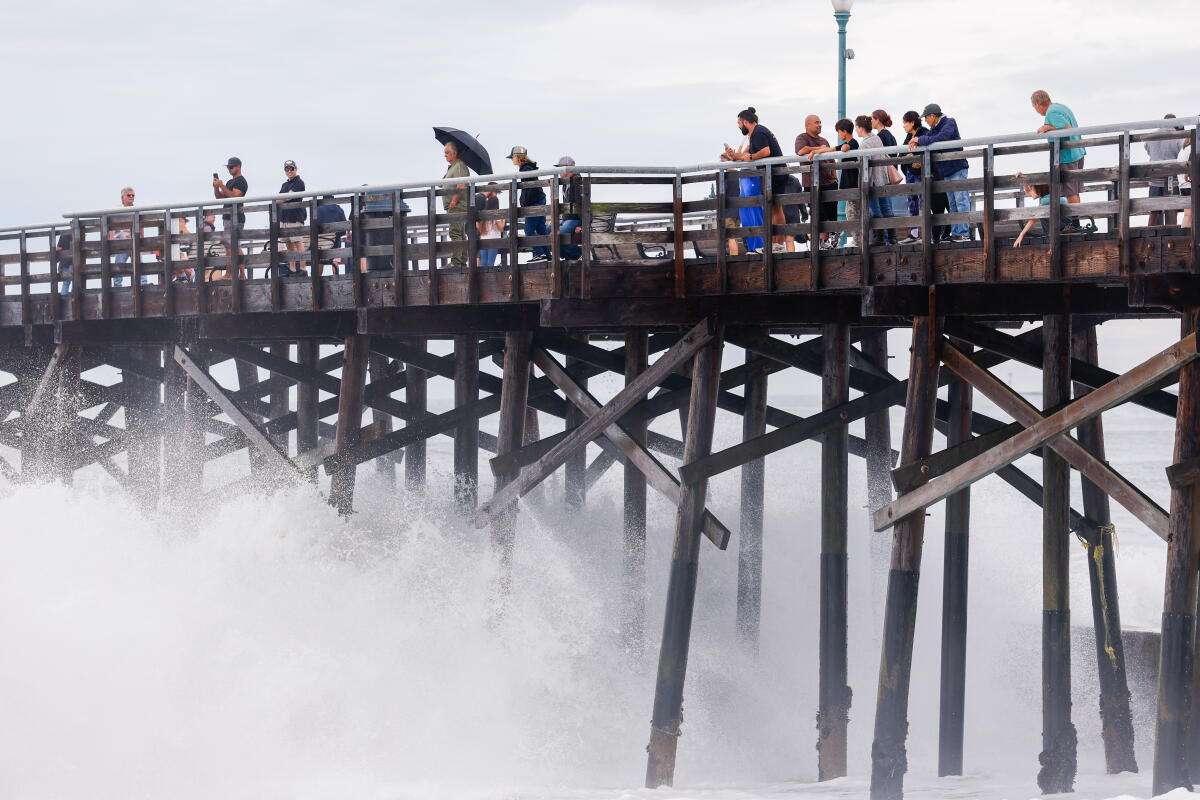 Waves crash into a wooden pier, with people on the pier.