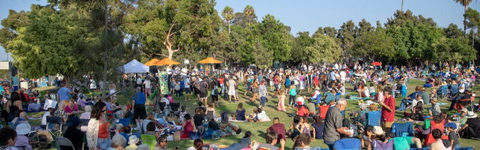 A large group of people stand and sit on a green lawn with umbrellas and picnic blankets