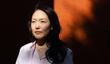 A woman with long black hair, wearing a light pink shirt and a circular necklace, stands against a dark orange background with shadows on her face.