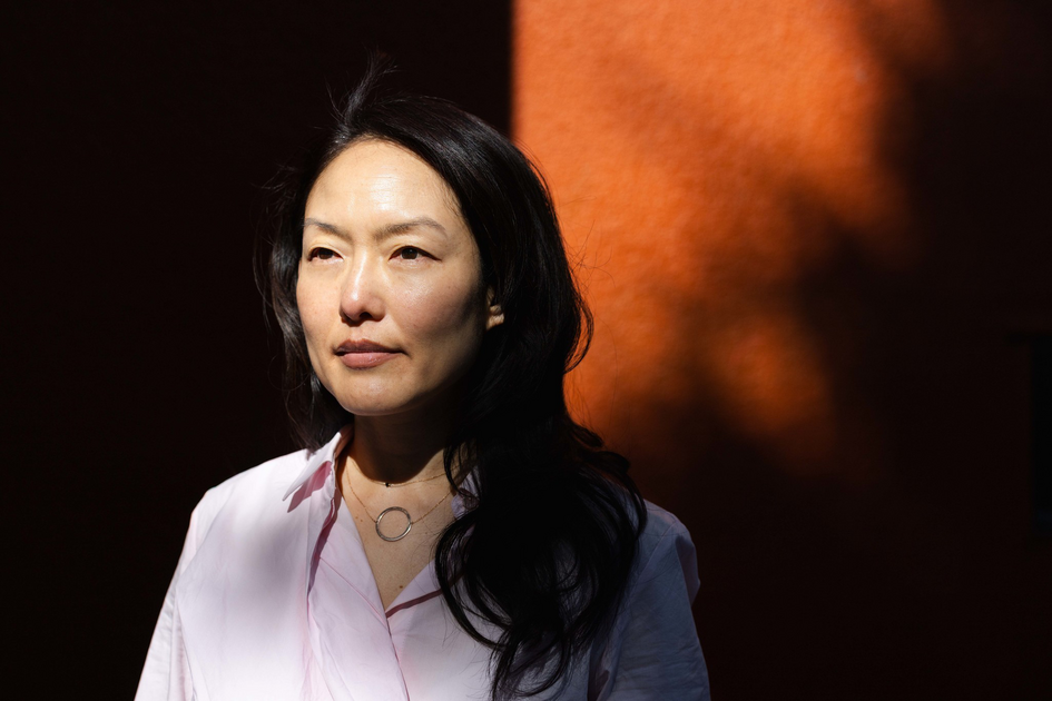 A woman with long black hair, wearing a light pink shirt and a circular necklace, stands against a dark orange background with shadows on her face.