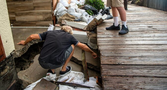 A person crouches down inspecting a collapsed concrete pathway leading to a home next to a boardwalk as another person, who's legs are only visible, watches over.