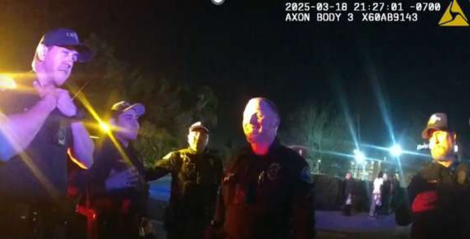 Police officers in uniform stand together at night, illuminated by bright lights with a crowd visible in the background.