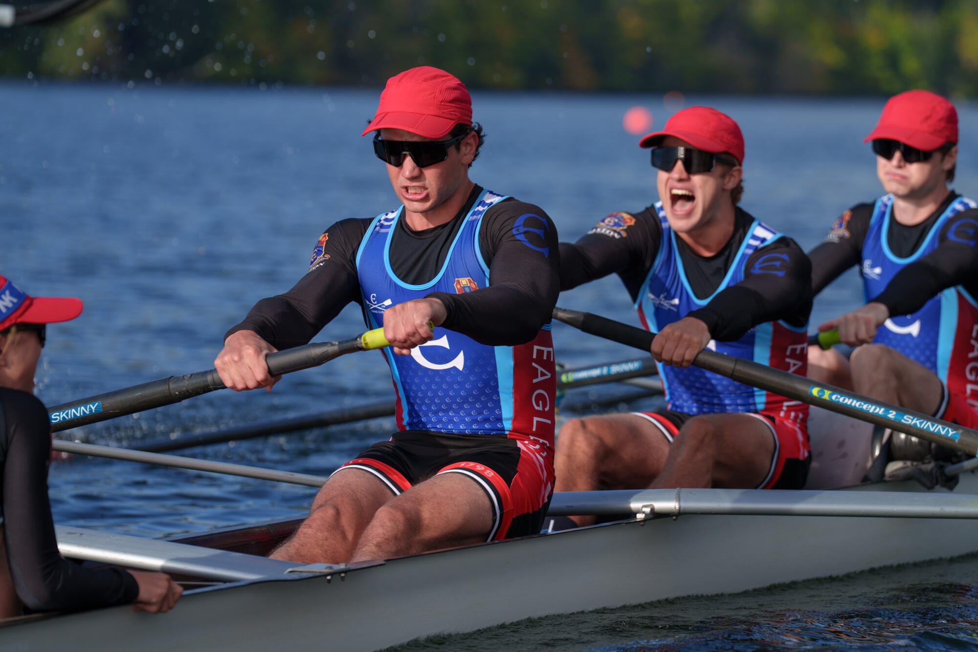 Three rowers in red ball caps and blue, red and black uniforms holding oars on a boat on the water. 