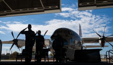 Two military personnel in silhouette are rendering a salute inside a hangar, with a large propeller-driven aircraft visible in the background against a partly cloudy sky. The scene appears to be a formal military ceremony or commemoration, likely involving the aircraft on display. The strong backlit composition creates dramatic silhouettes against the bright exterior.