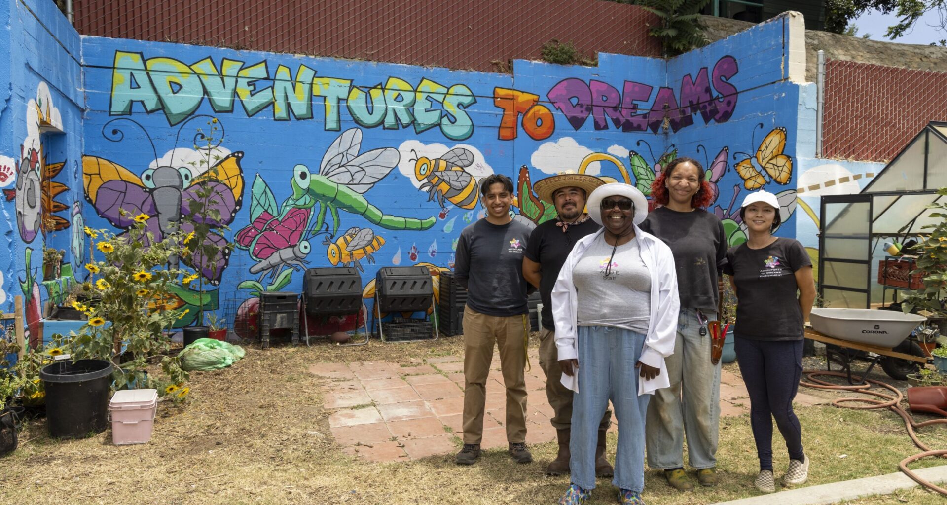 Five people stand smiling in front of a colorful mural that reads “Adventures to Dreams,” featuring cartoon insects and flowers, next to gardening equipment, sunflowers, and tables offering free food in an outdoor space.