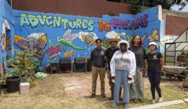 Five people stand smiling in front of a colorful mural that reads “Adventures to Dreams,” featuring cartoon insects and flowers, next to gardening equipment, sunflowers, and tables offering free food in an outdoor space.