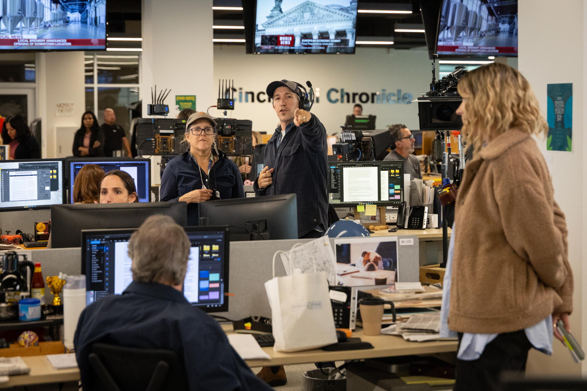 A group of people inside a newsroom under a row of TV sets