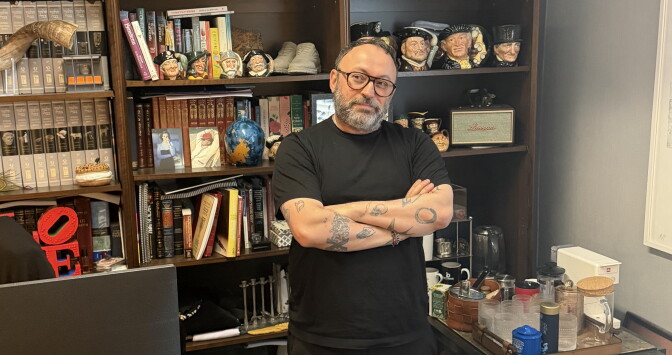 A bearded man with medium-light skin tone and glasses stands with his tattooed arms folded in an office lined with bookshelves.