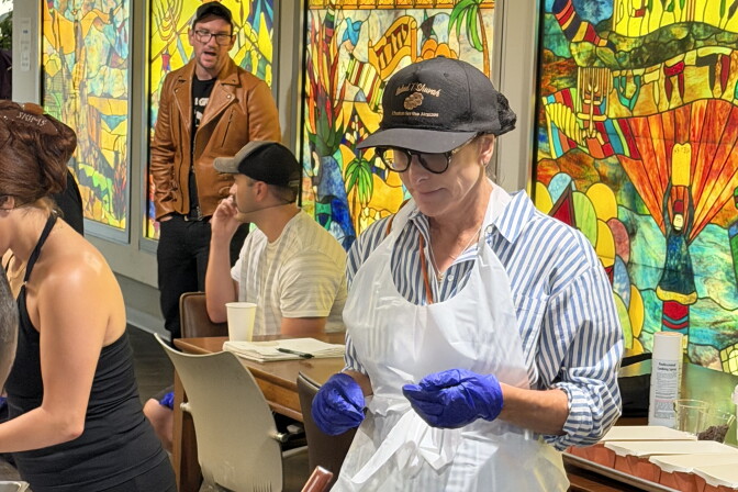 A woman with light skin tone — wearing a striped coat, a hat and purple latex gloves — looks at a table where baked goods are being prepared.