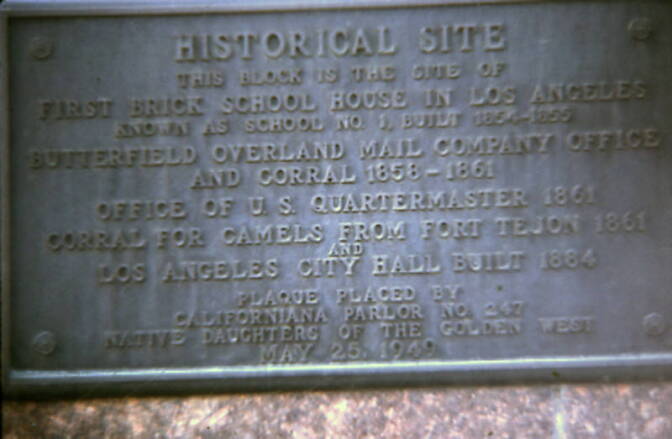 A close up of a gray plaque that reads: Historical Site. This block is the site of first brick school house in Los Angeles, known as School Number 1, built from 1854 to 1855. Butterfield Overland Mail Company Office and Corral, from 1858 to 1861. Office of the U.S. Quartermaster in 1861. Corral for camels from Fort Tejon in 1861. Los Angeles City Hall built in 1884. Plaque placed by Californiana Parlor Number 247, Native Daughters of the Golden West on May 25, 1949.