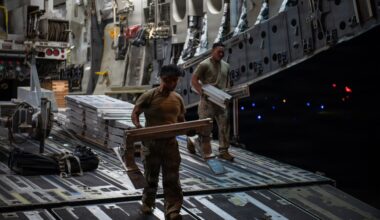 Two military personnel in combat uniforms are loading or unloading cargo inside what appears to be a large military transport aircraft, likely a C-17 Globemaster III. They are handling flat, aluminum-framed equipment cases or panels on the aircraft