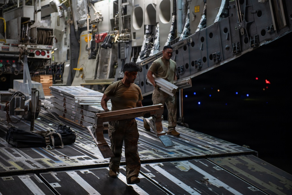 Two military personnel in combat uniforms are loading or unloading cargo inside what appears to be a large military transport aircraft, likely a C-17 Globemaster III. They are handling flat, aluminum-framed equipment cases or panels on the aircraft