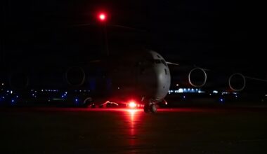 A large military transport aircraft, consistent with a C-17 Globemaster III, is photographed on a tarmac at night. The aircraft is illuminated primarily by its own red navigation/anti-collision lights, which cast a red glow on the ground beneath it. The dark background reveals distant airfield lighting along the horizon.