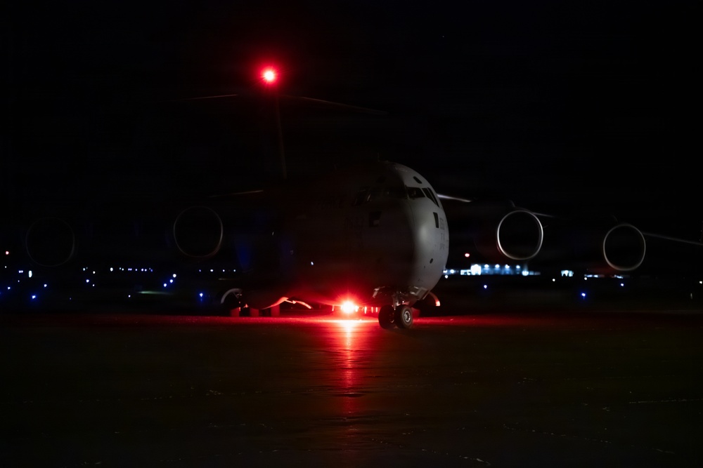 A large military transport aircraft, consistent with a C-17 Globemaster III, is photographed on a tarmac at night. The aircraft is illuminated primarily by its own red navigation/anti-collision lights, which cast a red glow on the ground beneath it. The dark background reveals distant airfield lighting along the horizon.
