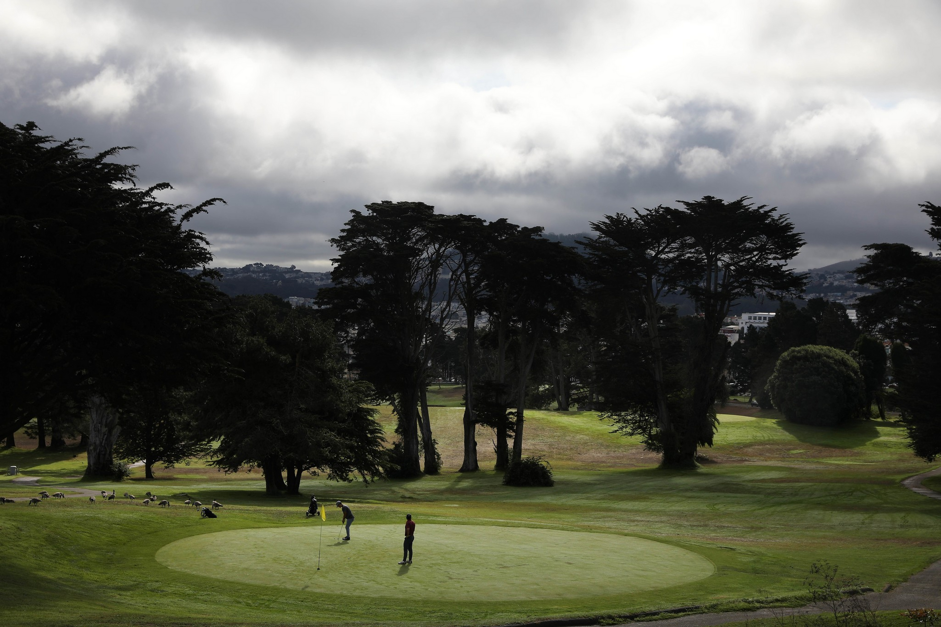 Two people play golf on a green surrounded by tall trees under a cloudy sky with hills and buildings in the distant background.