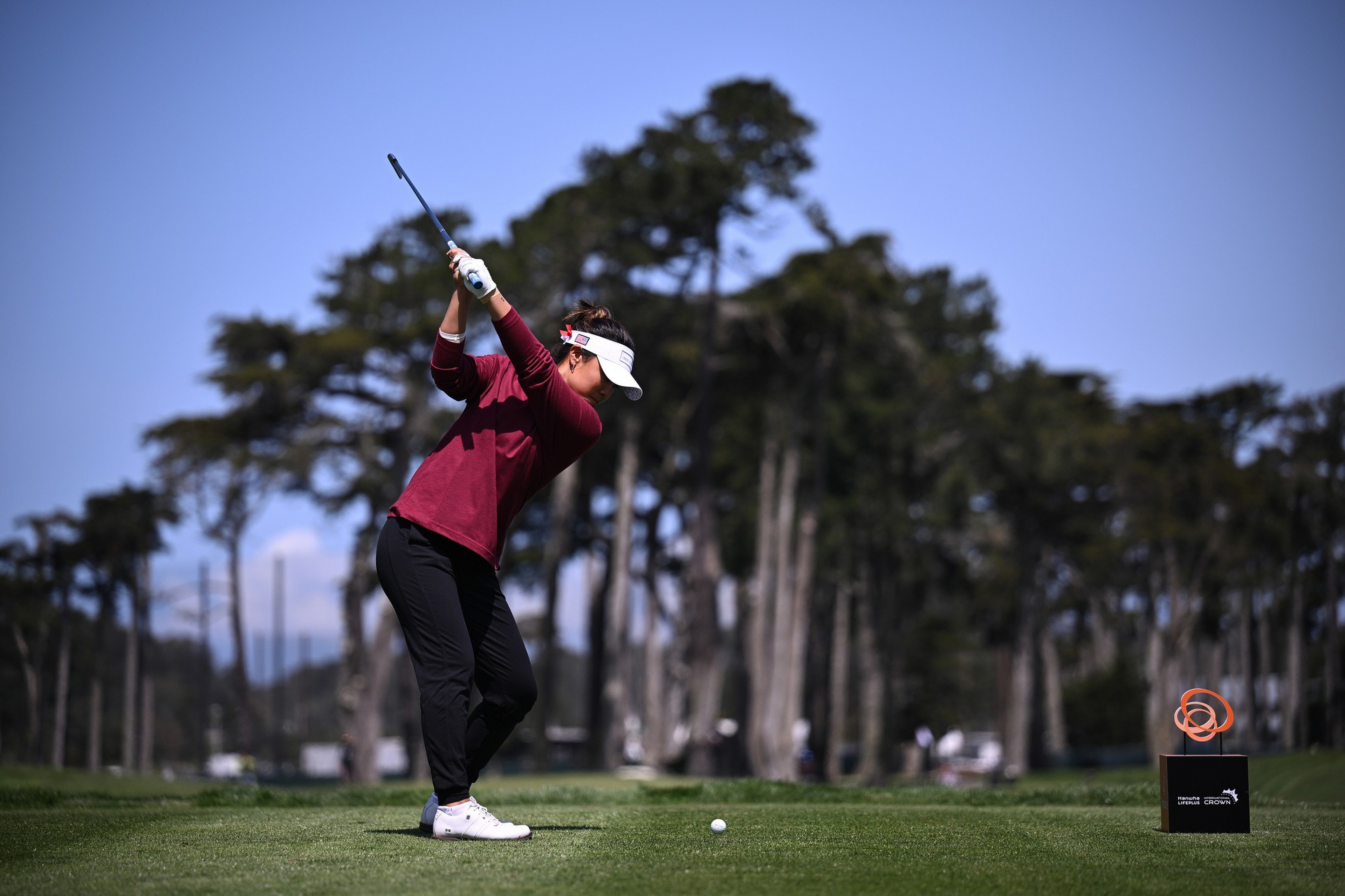 A golfer in a maroon top, black pants, and white visor is mid-swing on a grassy course with tall trees in the background under a clear blue sky.