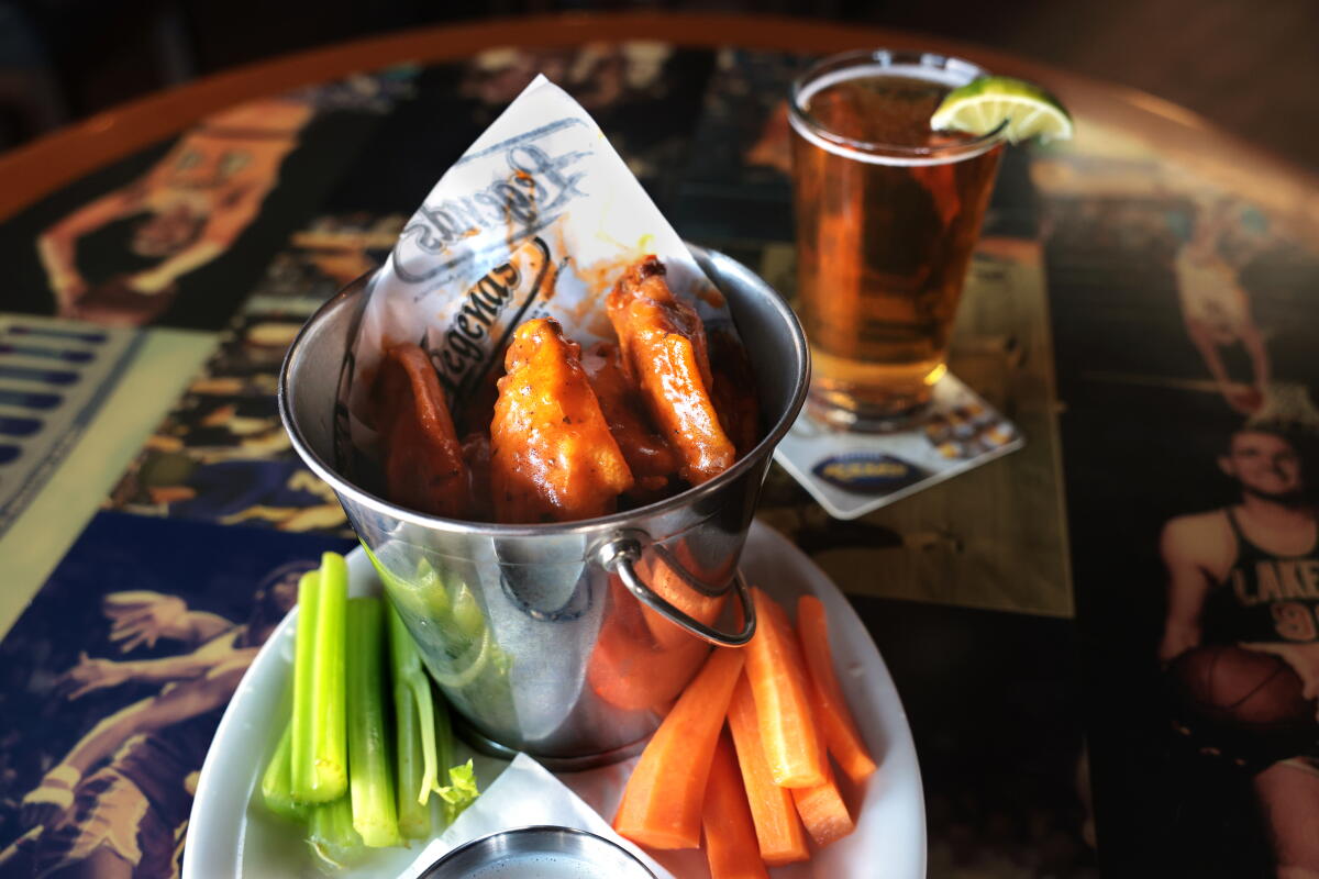 A small metal pail of chicken wings on a plate with sliced celery and carrots; nearby, a pint glass of beer with a lime wedge